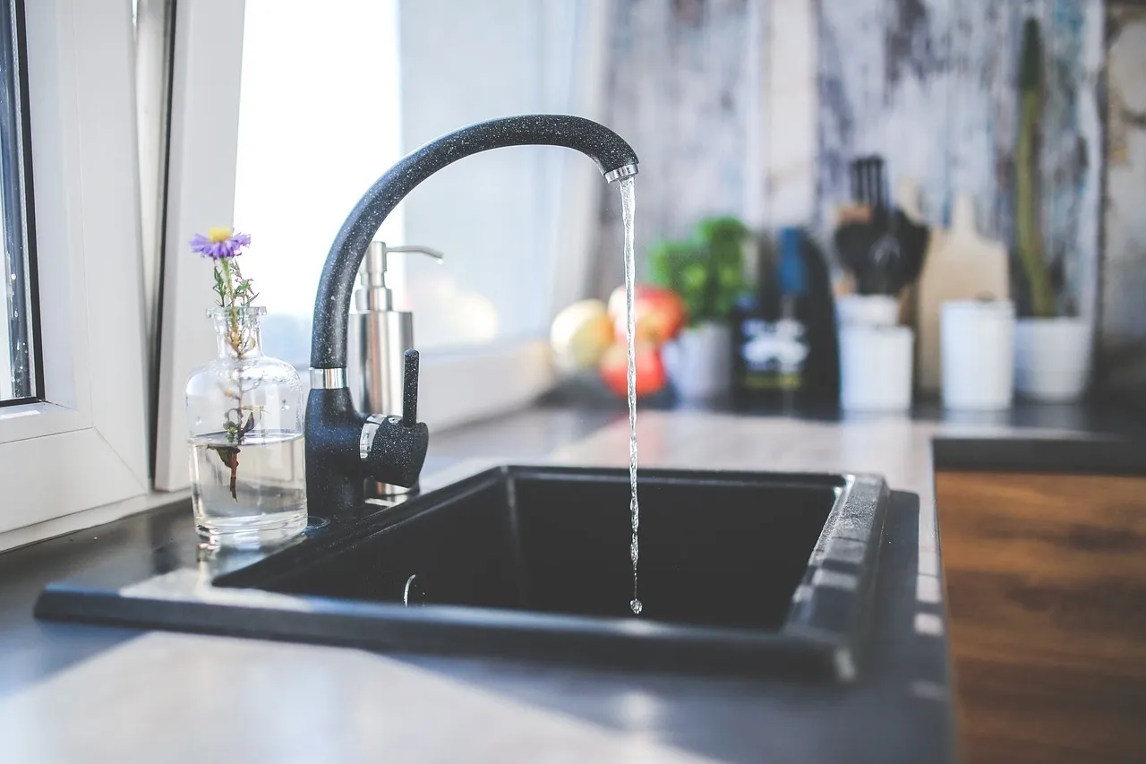 Modern black kitchen faucet with running water over a black sink next to a window and a small flower vase.