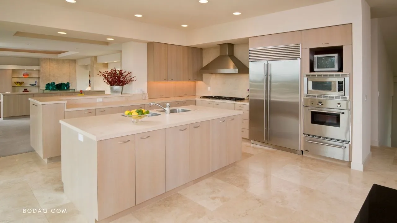 Spacious modern kitchen with light wood cabinetry, a large island with sink, stainless steel appliances, and beige tiled floor.