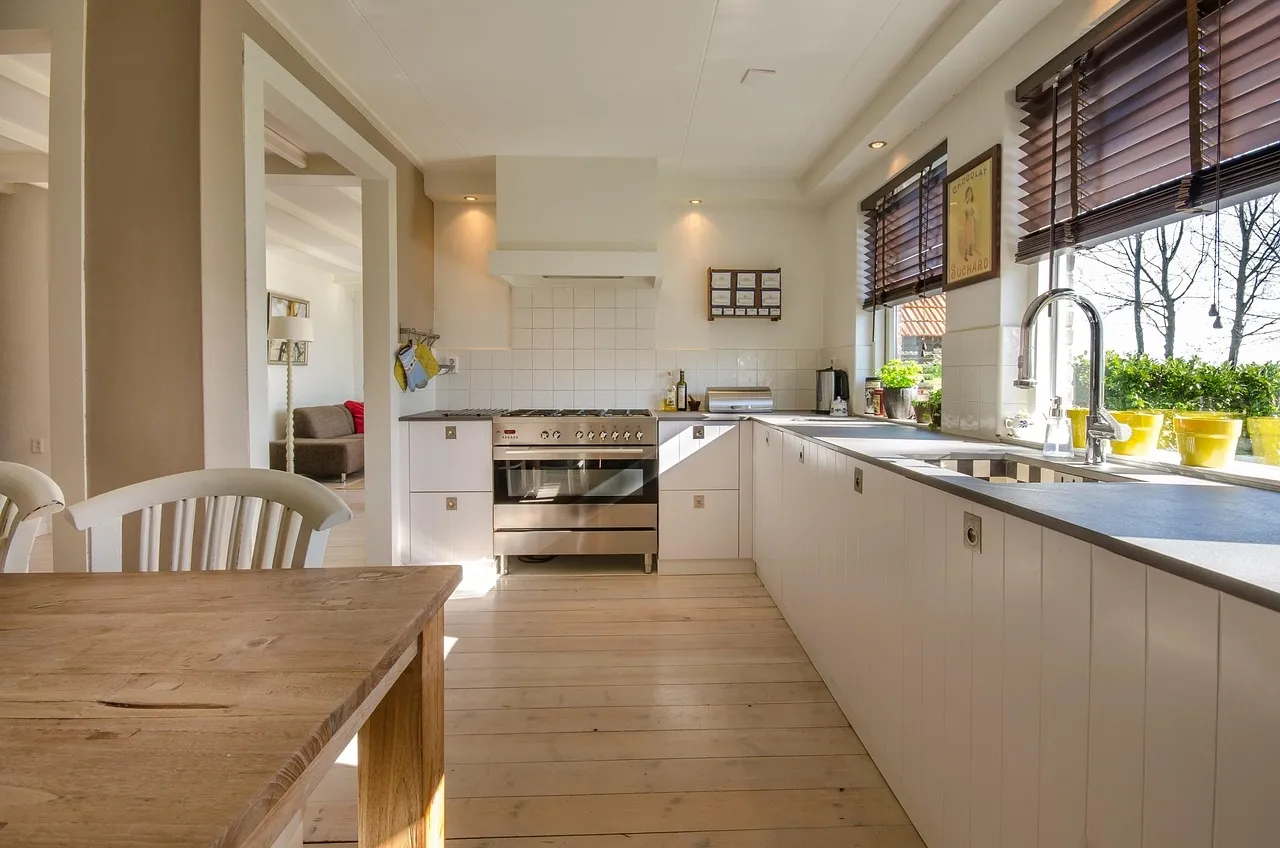 Bright modern kitchen with white cabinets, stainless steel stove, wooden table, and large windows with brown blinds.